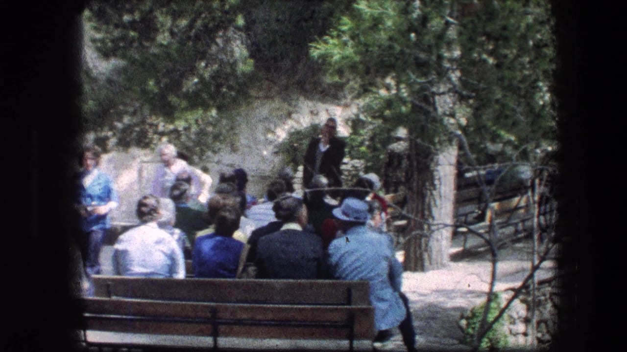 Wide view of adults seated on benches listening to a speaker in a garden area.