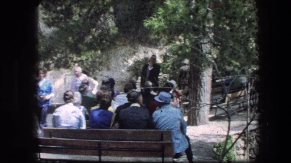 Wide 1976 Garden Tomb view with adults on wooden benches listening to a speaker beside trees, railings, stonework, and sunlit paths.