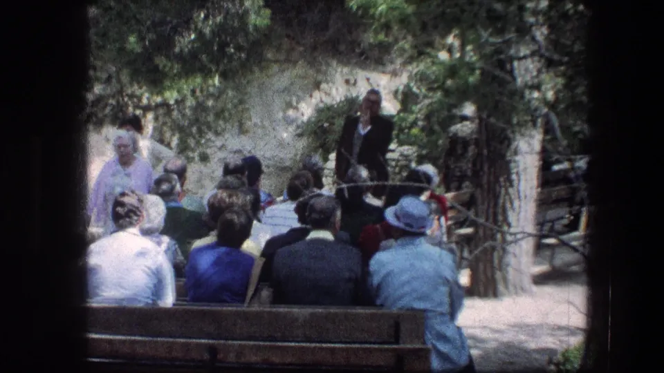 1976 8mm frame from Jerusalem showing the audience from behind as they face a speaker in the Garden Tomb's outdoor setting.
