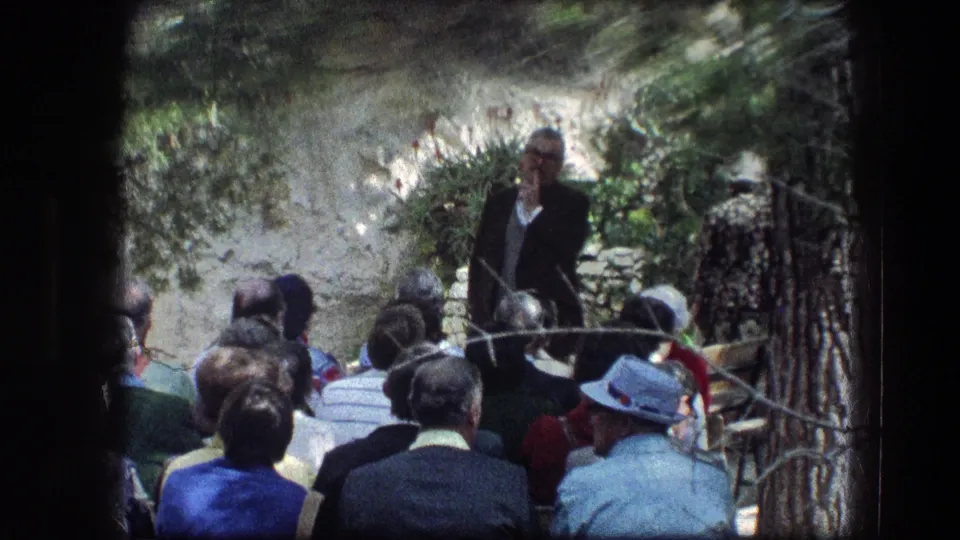Close 1976 Garden Tomb view of a suited man speaking with a microphone while adults sit in rows beneath trees and beside stonework.