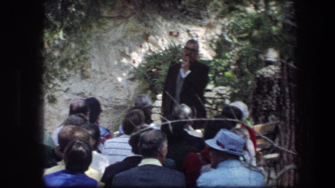 Tour guide speaking to a seated group in a garden setting.