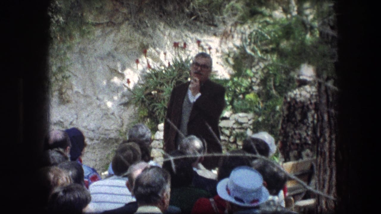 A man in a dark suit stands before a tour group at the Garden Tomb, with stonework and plants behind him.