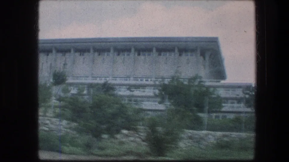 1976 8mm view in Jerusalem showing a broad modern building behind trees and shrubs, softened by vehicle motion.