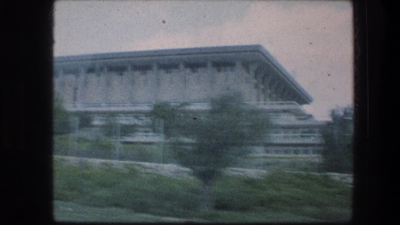 A moving 8mm frame shows the corner of a large Jerusalem building with landscaped greenery in the foreground.