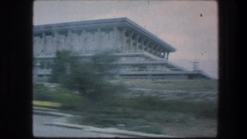 Vehicle-motion blur softens a side view of the 1976 Jerusalem building, with low greenery and roadway edge visible.