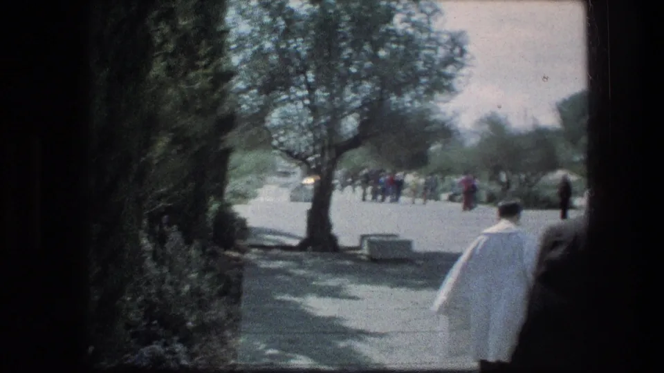A Jerusalem walkway scene shows trees, distant visitors, and a man in a white coat near the right edge of the 8mm frame.