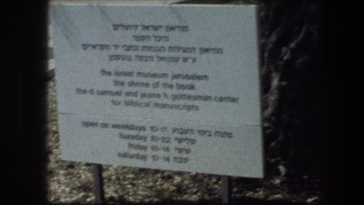 Grey bilingual sign standing outdoors with dark foliage and stone ground behind it.