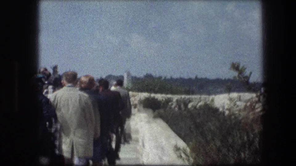 Visitors move along a pale stone walkway in Jerusalem-area 1976 footage, with low walls, shrubs, and distant landscape visible.