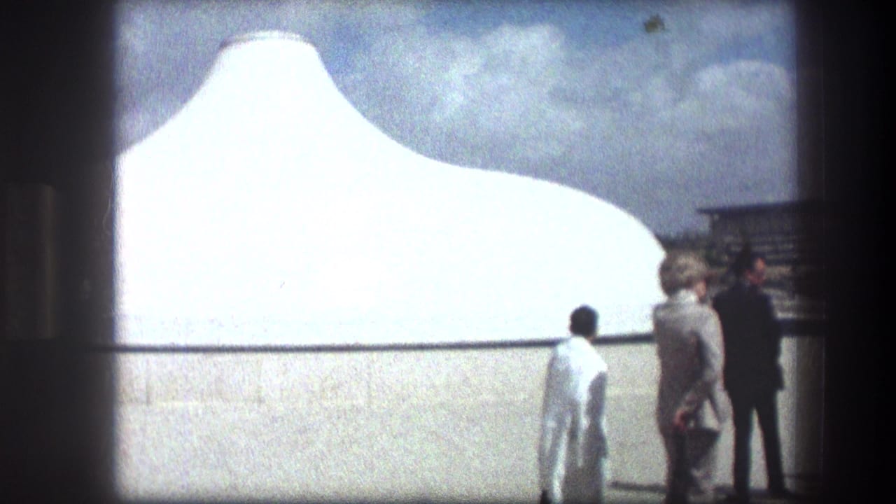 Visitors stand near a white curved tomb structure under a bright sky.
