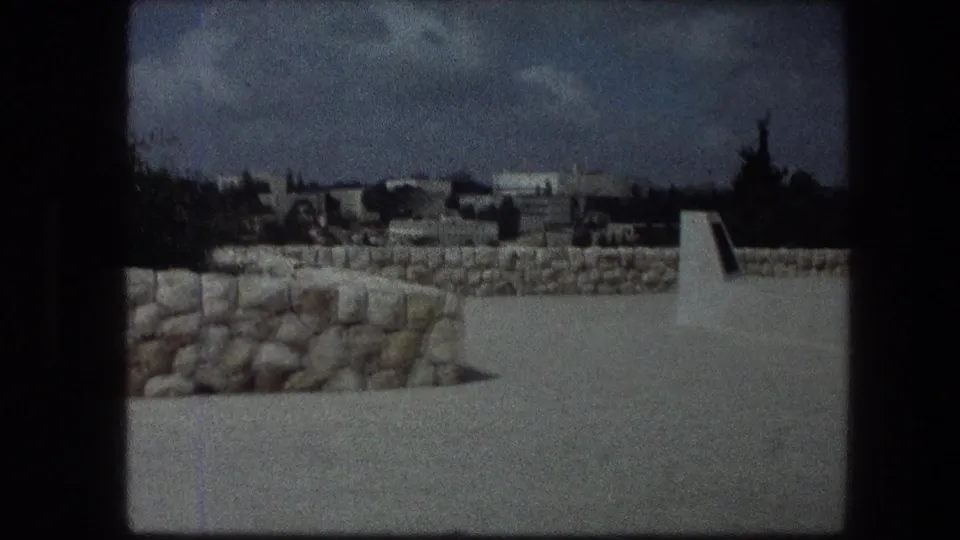 The Garden Tomb grounds show a stone wall, pale walkway, trees, and a distant Jerusalem view.