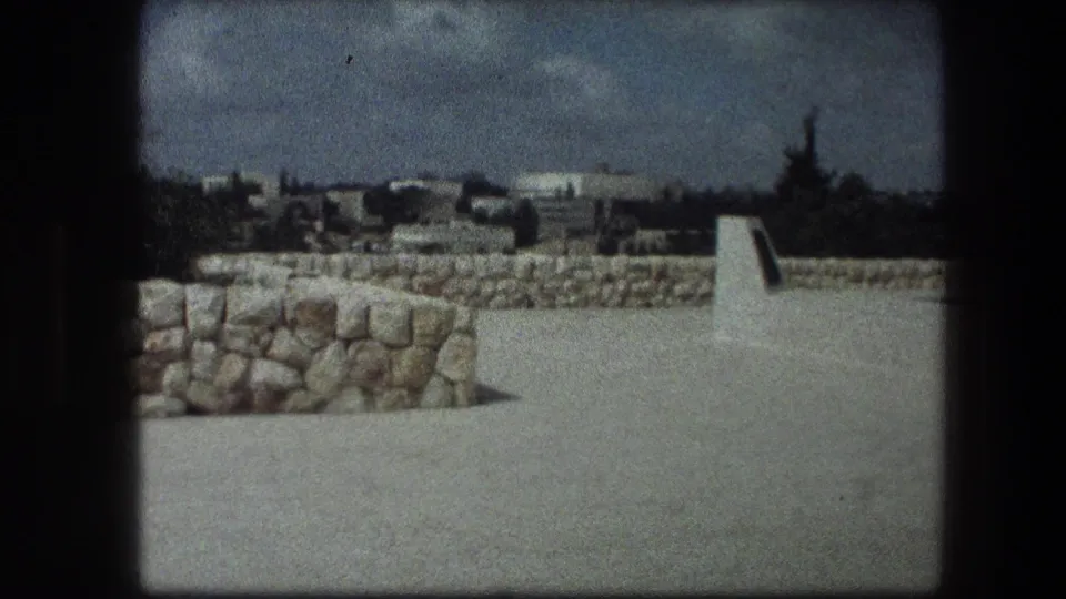 Garden Tomb grounds in Jerusalem seen in a grainy 1976 home-movie frame, with low stone walls, open paving, trees, and distant structures.