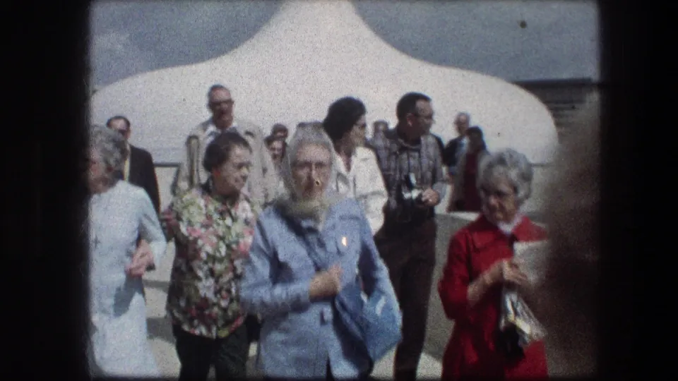 Adult tourists in 1970s clothing walk near a white structure at the Garden Tomb in Jerusalem, filmed on 8mm.