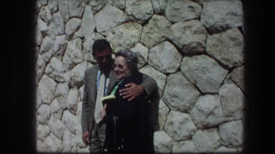 In 1976 Jerusalem home-movie footage, an older woman smiles while a man in a suit holds her close near a stone wall.