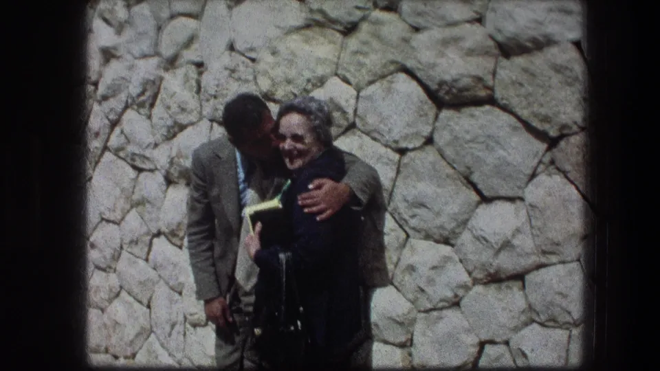 In 1976 Jerusalem footage, a young couple stands close together before a rough pale stone wall, with the man leaning toward the smiling woman.