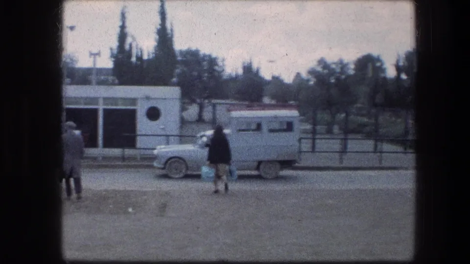 1976 Jerusalem footage shows a pale truck beside a small white building with a circular window while pedestrians move through the foreground.