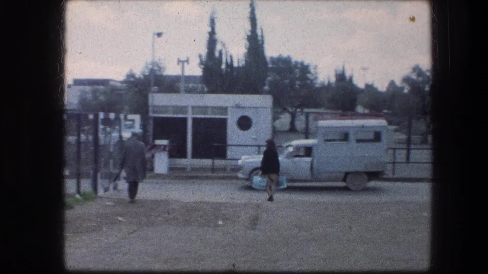 At the Garden Tomb area in Jerusalem, pedestrians pass a white roadside building and a pale truck under an overcast sky.