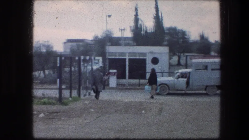 1976 8mm view near the Garden Tomb in Jerusalem, showing visitors by a gate, a small white structure, and a parked car.