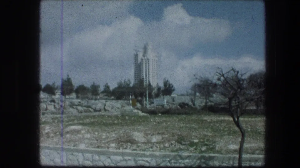 1976 Jerusalem view from the Garden Tomb area, showing grass, low stone ground, trees, and a tall white building in the distance.