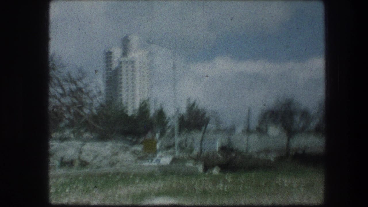 Tall white building beyond rocky roadside and sparse trees, seen through a dark 8mm frame border.