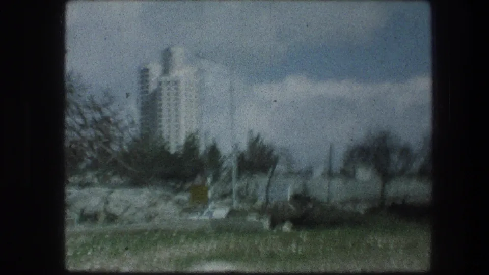 1976 Jerusalem-area home-movie frame showing a distant white high-rise beyond rocks, trees, and roadside blur.