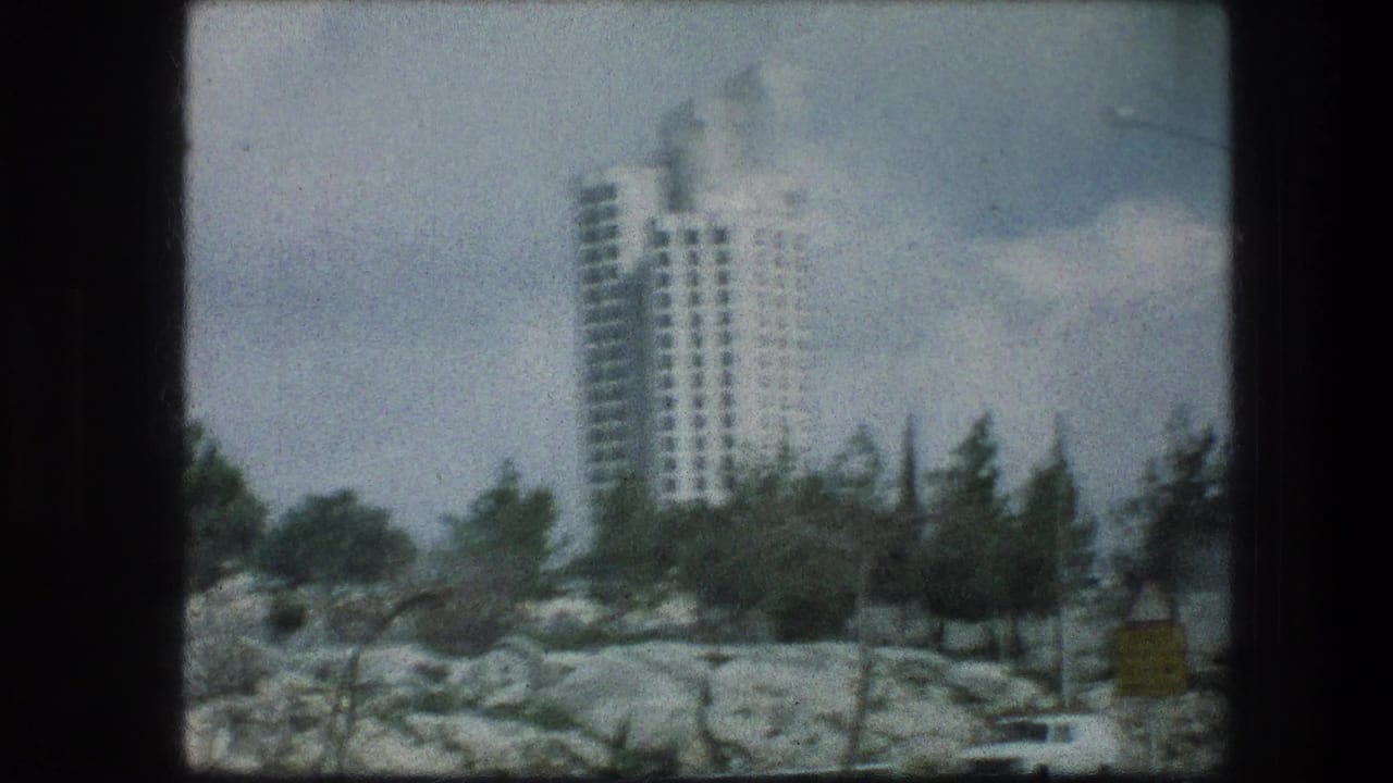 White high-rise behind rocky ground and trees under a cloudy sky.
