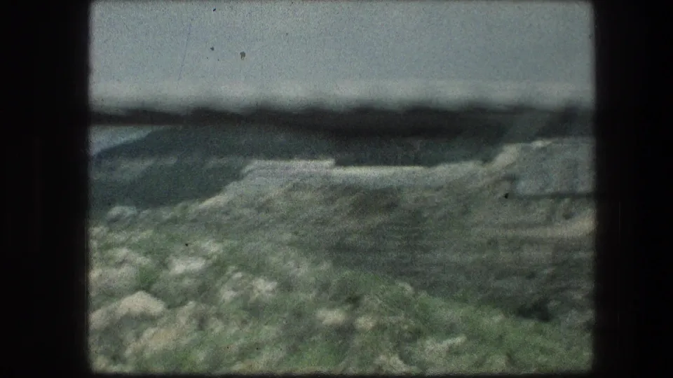 Handheld 8mm frame near the Garden Tomb area, looking across rocks, dry grass, and a low Jerusalem hill under hazy light.