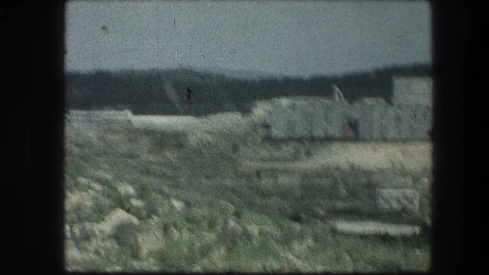 1976 8mm frame near the Garden Tomb showing rocky ground, sparse vegetation, distant ridge, and a pale hillside structure.