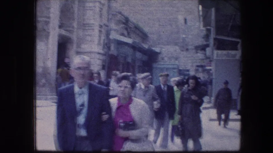 1976 home-movie view of adults in suits, coats, and dresses walking along a narrow Jerusalem stone street.