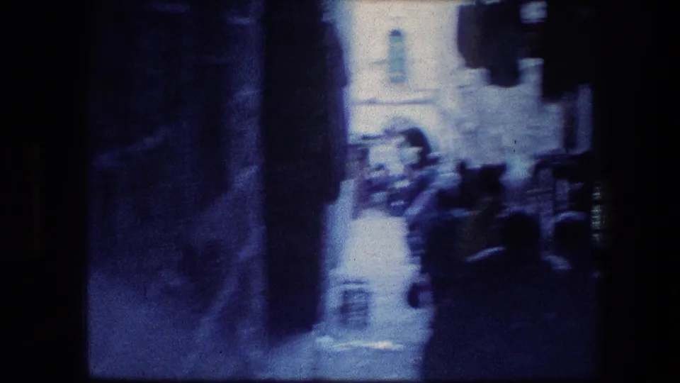 A dim handheld street view looks from shade toward a brighter Jerusalem passage with pedestrians ahead.