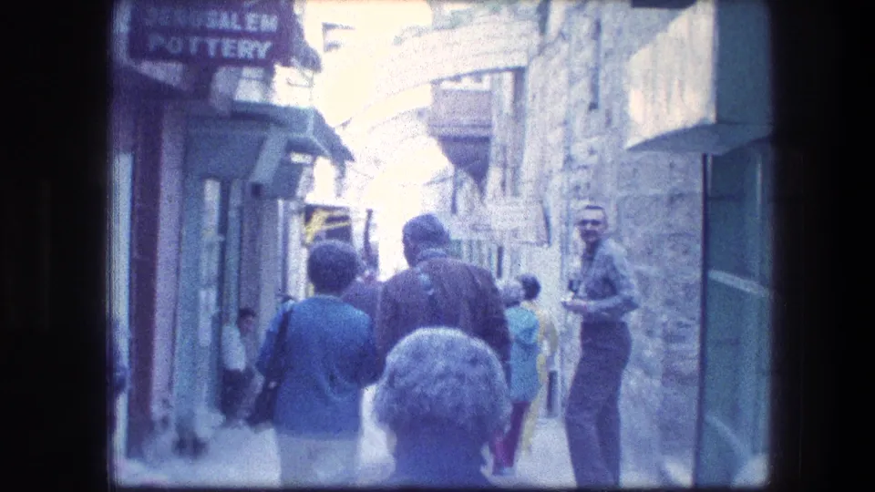 Visitor-shot 1976 8mm frame of people walking away through a Jerusalem stone passage, with partial shop signage and bright sun ahead.