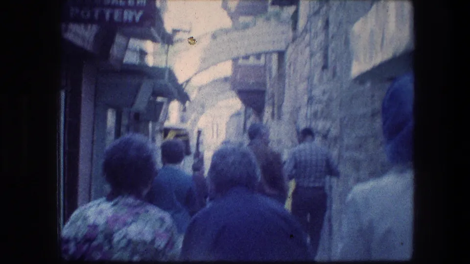 Soft, blue-tinted 8mm frame of a crowded Jerusalem alley near the Garden Tomb, with pedestrians moving away under stone arches.