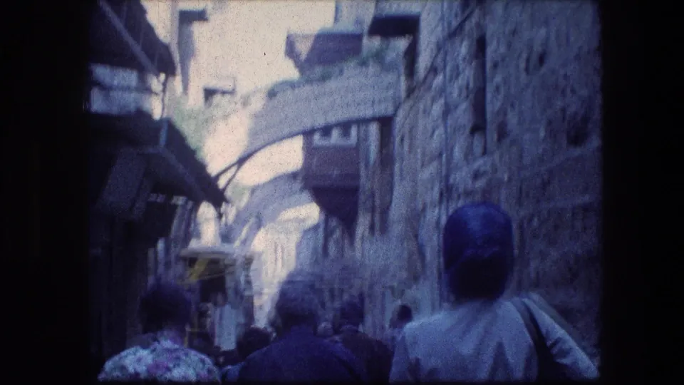 Handheld 8mm frame of Jerusalem visitors walking through a tight stone passage, with aged color and motion blur.