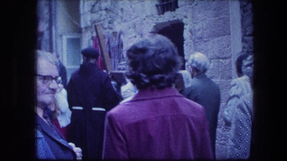 1976 Jerusalem home-movie frame showing older visitors and a robed participant with a wooden cross in a narrow stone passage.