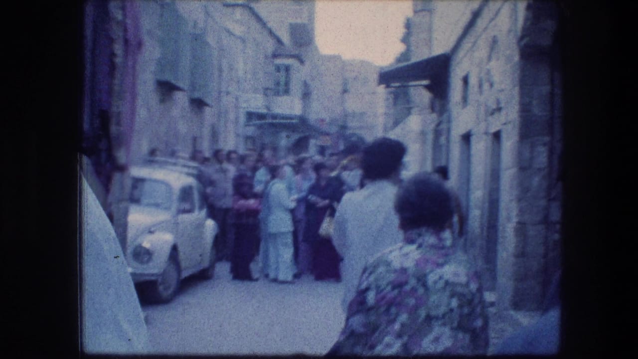1976 8mm view of a crowded narrow street near the Garden Tomb in Jerusalem, with stone buildings and a white car at left.
