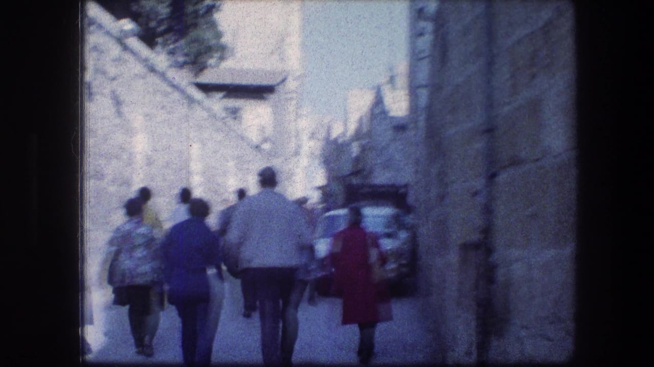 Several pedestrians walk along a sunlit stone lane with a parked car at right.