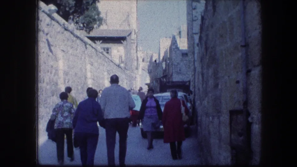 Handheld 8mm view of tourists walking through a narrow Jerusalem stone street, with parked cars and bright daylight visible.