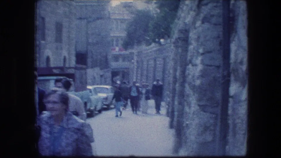 Wider 1976 home-movie view down a Jerusalem stone street with pedestrians, parked cars, and distant buildings.