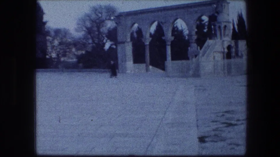 1976 Garden Tomb Jerusalem frame showing a quiet paved courtyard, arched stone openings, trees, and a distant visitor.