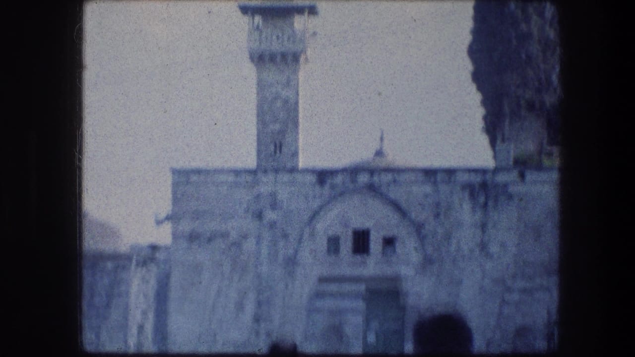 1976 8mm Jerusalem frame showing pale stone architecture, an arched facade and a tall minaret-like tower near dark cypress trees.