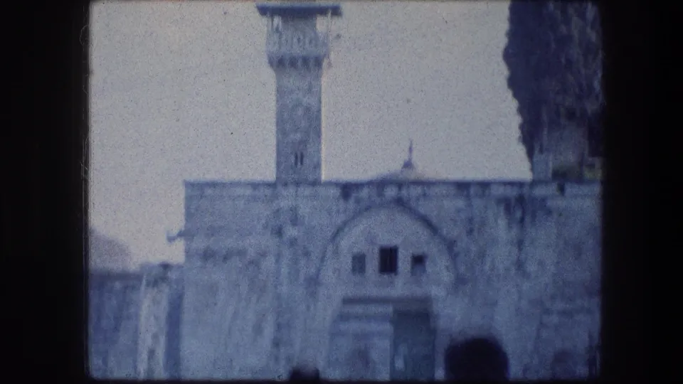 1976 8mm Jerusalem frame showing pale stone architecture, an arched facade and a tall minaret-like tower near dark cypress trees.
