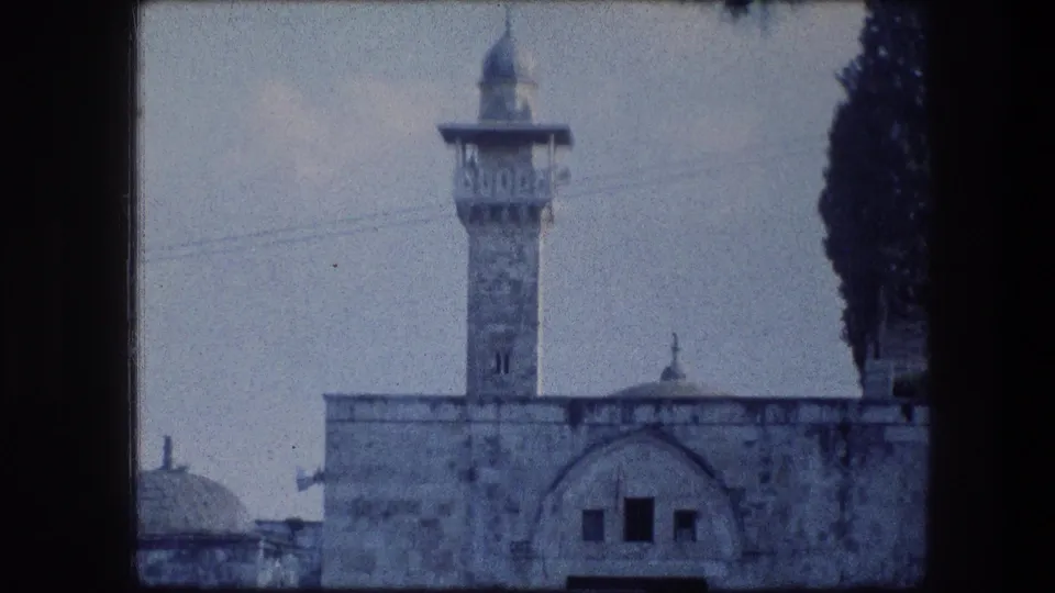 1976 8mm Jerusalem view of a minaret-like tower rising over stone walls, with a dome partly visible and a dark tree at right.