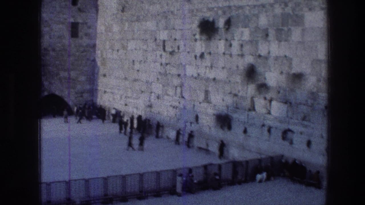 Visitors stand near a large stone wall behind metal barriers in a Jerusalem religious courtyard.