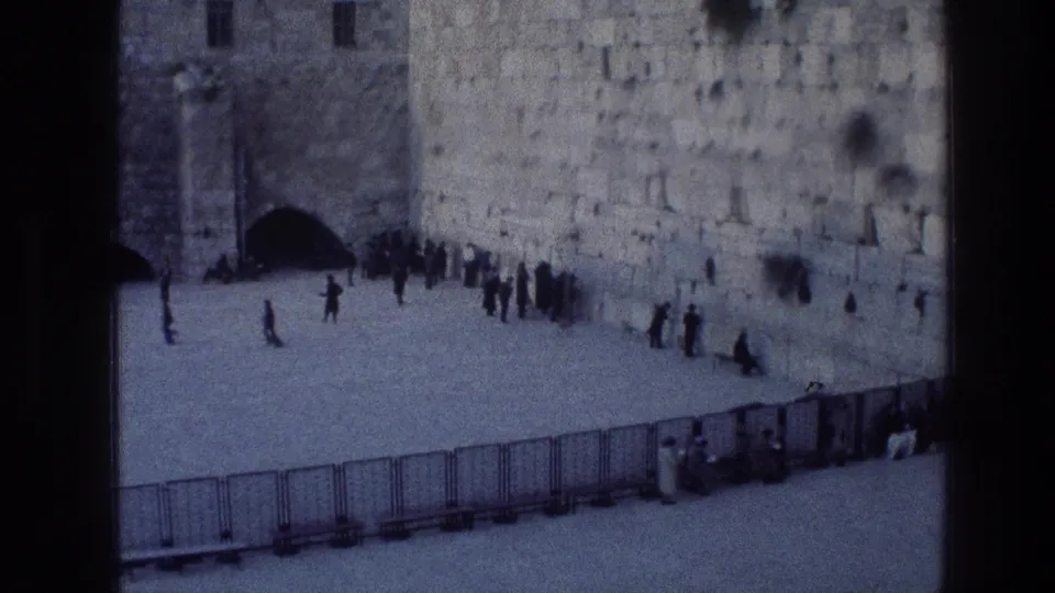 Wider 1976 8mm view of a Jerusalem religious site, with an arched entrance, stone wall, barriers, and visitors gathered in prayerful stillness.