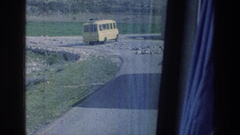 1976 Israel frame with a yellow bus rounding a rural bend, seen past window reflections and a dark curtain edge.