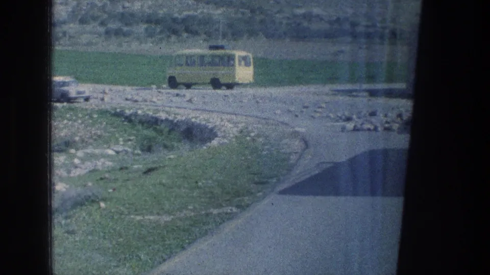 A yellow van and a pale car appear on a rural road in Israel, with fields, rocky ground, and film-frame borders visible.