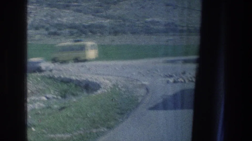 A blurred yellow van and pale car move through a rural road scene in 1970s Israel, with fields and rocky terrain in view.