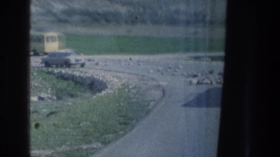 A roadside view from 1976 archival home-movie footage shows a pale car and yellow bus beside rocks, grass, and a low hillside.