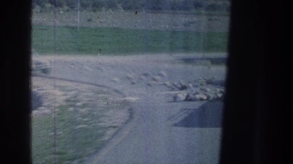Soft 8mm frame showing a rocky roadside, green slope, and part of a pale vehicle at the left edge during a moving travel shot.