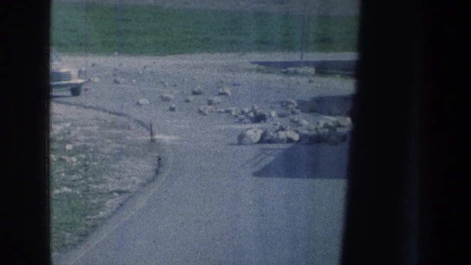 A 1976 8mm travel frame shows a curving paved road, scattered stones, grass, and the front of a light-colored car at the edge.