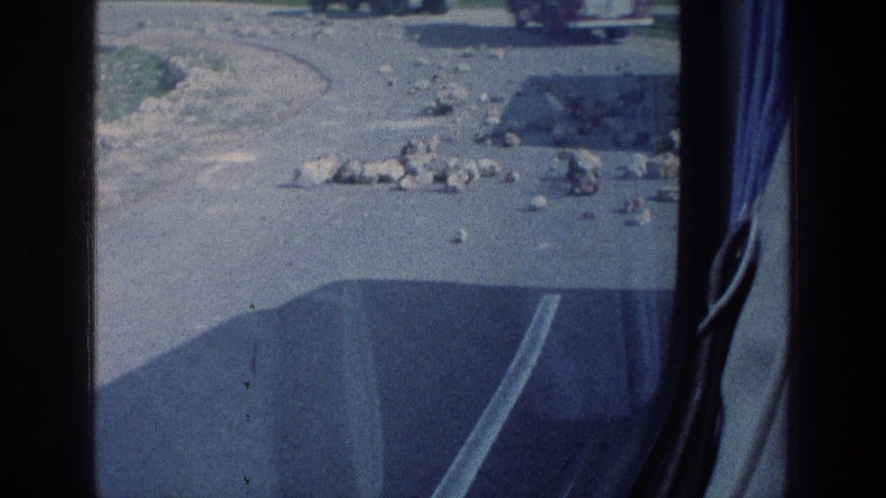 1976 8mm travel view from inside a vehicle, showing scattered stones on a paved road and distant vehicles beyond the window frame.